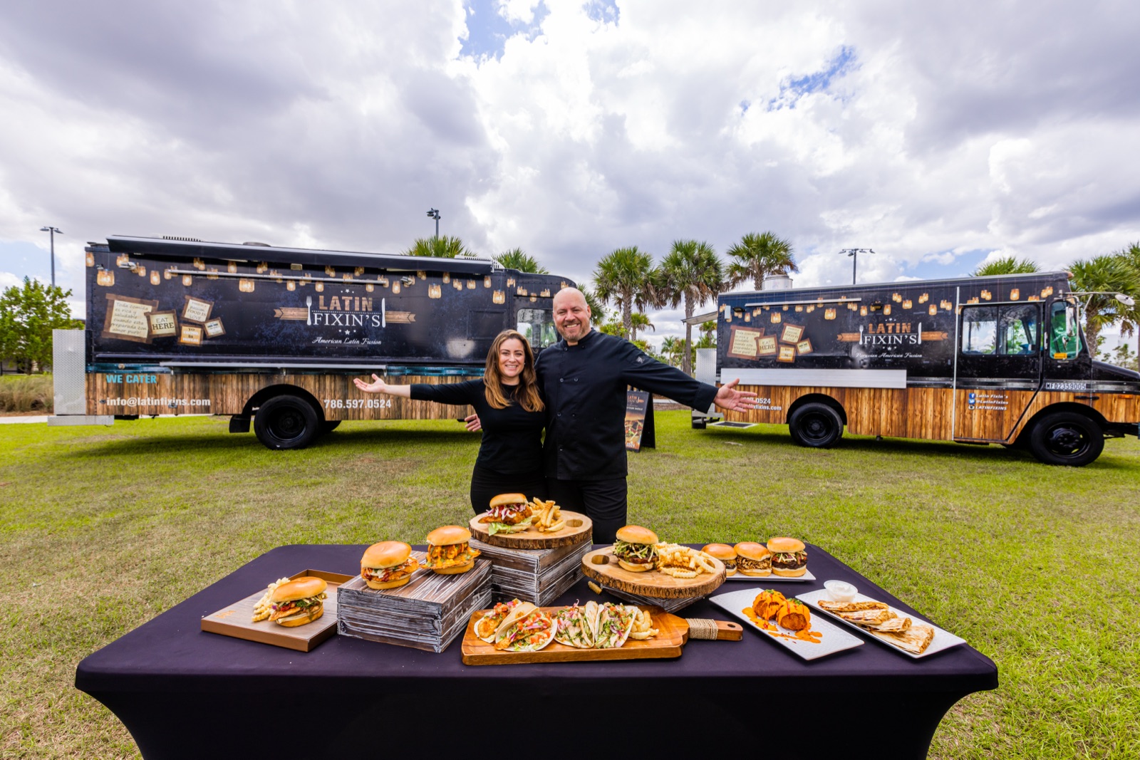 Chef Alex and Kiara standing between two Latin Fixin's food trucks with a full spread of food on the table in front of them